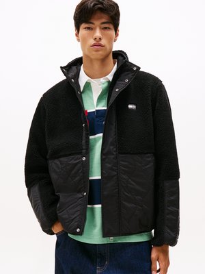 Black fleece and matte nylon jacket with snap closure, front pockets, and quilted texture. Worn over a multicolored striped polo shirt.