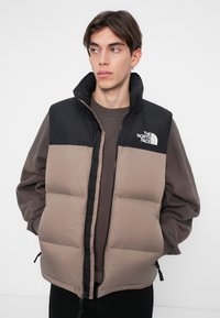 Mīksts veste melnā un brūnā krāsā, ar augstu apkakli, rāvējslēdzēja priekšpusi, sānu auklām un The North Face logo augšējā kreisajā pusē.