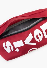 Saco vermelho com fecho de correr feito de tecido texturizado; apresenta um interior cinzento e letras brancas em destaque no exterior. Design compacto, forma cilíndrica.