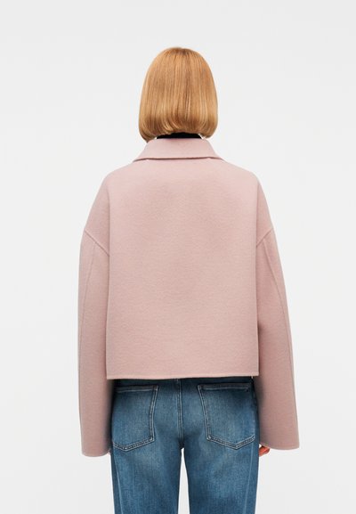 Άτομο με κοντά ξανθά μαλλιά που φοράει ένα ανοιχτό ροζ κοντό μπουφάν και μπλε τζιν, εμφανιζόμενο από πίσω έναν απλό φόντο.