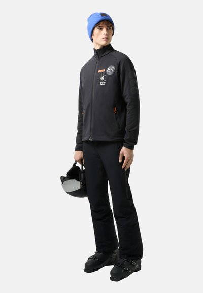 Black fleece jacket with orange zippers and patches, paired with black pants and a blue knit beanie. Holds a black ski helmet.