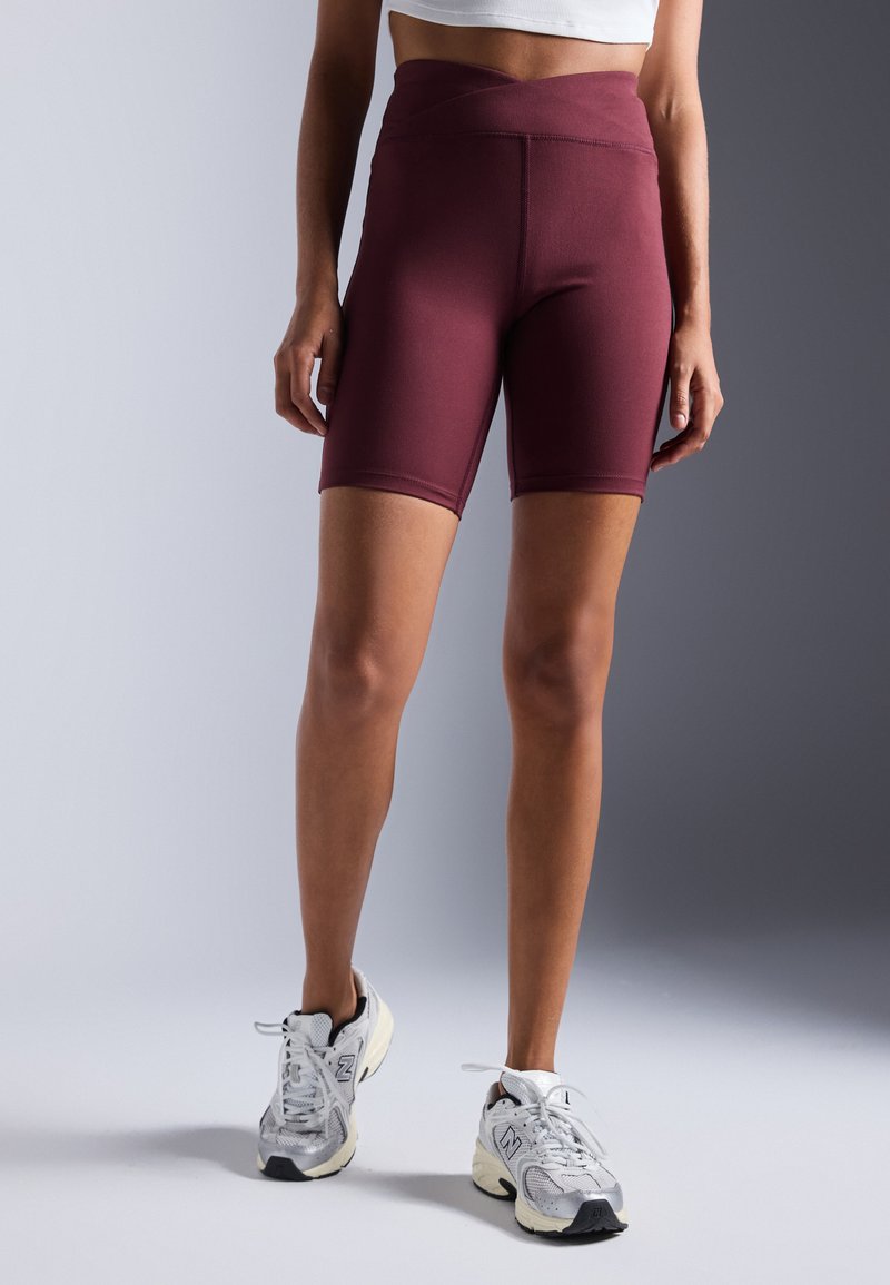 Burgunderfarbene, hochtaillierte Radshorts mit einer glatten Textur und einem überkreuzten Bund. Kombiniert mit grauen Sportschuhen.