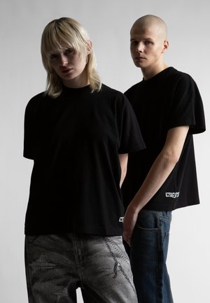 Dwóch młodych dorosłych ubranych w czarne, za duże T-shirty i dżinsy stoi na tle gładkiego, jasnego tła, jeden z krótkimi blond włosami, drugi z ogoloną głową.