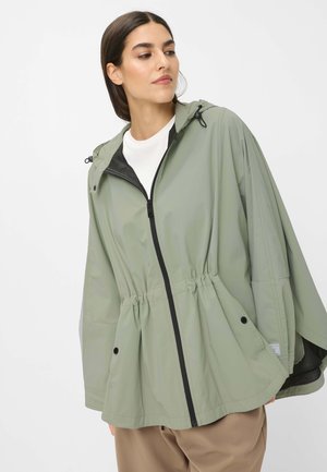 Frau trägt hellgrüne Kapuzen-Regenjacke mit schwarzem Reißverschluss und Kordelzug in der Taille, beige Hose, schaut seitlich vor weißem Hintergrund.