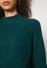 Een persoon met krullend haar, die gouden ringoorbellen draagt en een donkergroen geribbelde breiselsweater met een hoge hals.