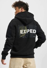 Svart hettegenser med et grafisk patch på ryggen som har teksten "EXPED" og abstrakte linjemønstre i gult og grønt.