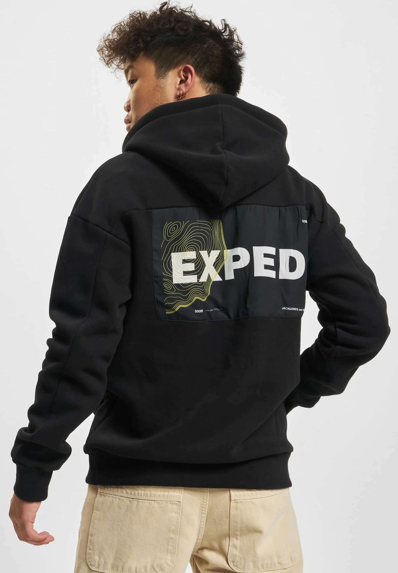 Svart hettegenser med et grafisk patch på ryggen som har teksten "EXPED" og abstrakte linjemønstre i gult og grønt.