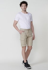Pantaloni scurți cargo bej cu multiple buzunare și talie cu șnur, asortați cu un tricou alb simplu și adidași negri. Textură netedă.