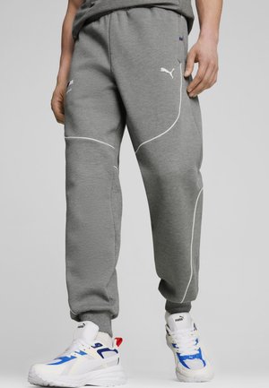 Person iført grå Puma joggingbukser og hvide sneakers med blå og røde detaljer står mod en enkel baggrund.