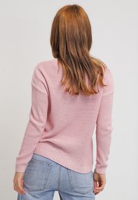 Pull tricoté rose clair à coupe décontractée, surface texturée et ourlet côtelé. Porté avec un jean en denim bleu clair et de longs cheveux bruns visibles.