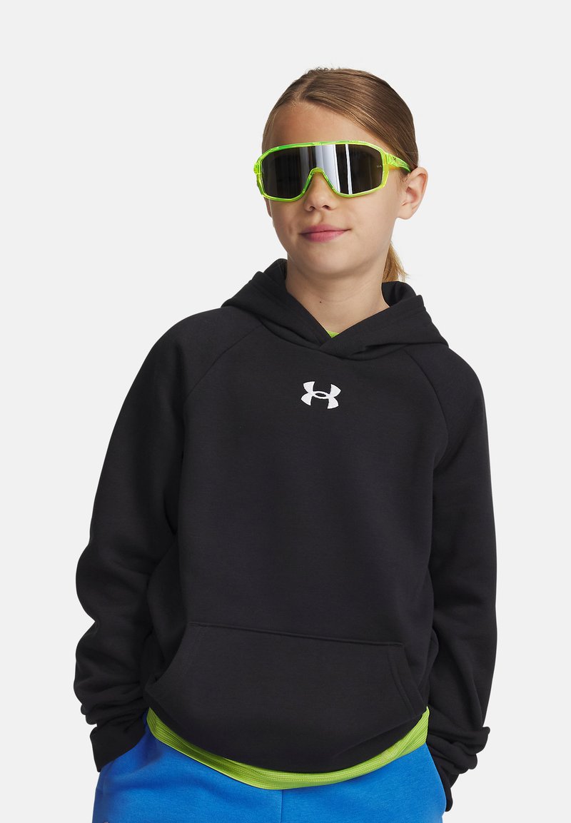 Jeune personne portant des lunettes de soleil réfléchissantes vert fluo, un hoodie noir Under Armour et un pantalon bleu, se tenant les mains dans les poches.