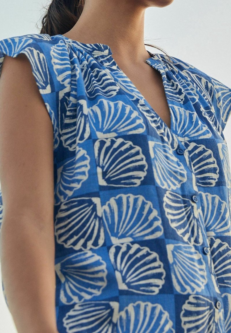 Chemise à boutons bleue avec un motif coquillage, dotée de courtes manches volantées et d'un décolleté en V, fabriquée en tissu léger.