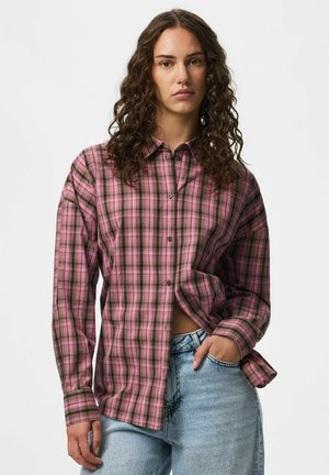 Femme aux cheveux bouclés portant une chemise à carreaux rose et marron boutonnée et un jean bleu clair, debout avec une main dans la poche.