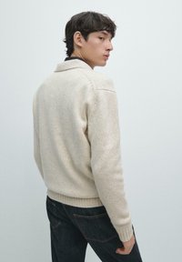 Kremowy sweter o dzianinowej fakturze, z prążkowanym wykończeniem na dole i mankietach oraz klasycznym kołnierzem. Noszony z ciemnymi dżinsami, widziany od tyłu.