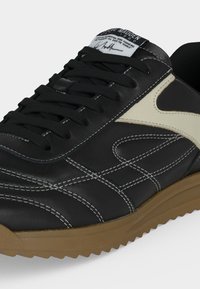 Μαύρο δερμάτινο αθλητικό παπούτσι με λευκή πλαϊνή ρίγα, μαύρα κορδόνια, λευκά ραφές και καφέ υφασμένη λαστιχένια σόλα.