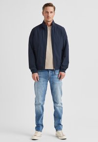 Marineblaue Bomberjacke mit Stehkragen und Reißverschlusstaschen, getragen über einem hellbeigen Hemd und hellblauen Jeans, komplettiert mit weißen Turnschuhen.