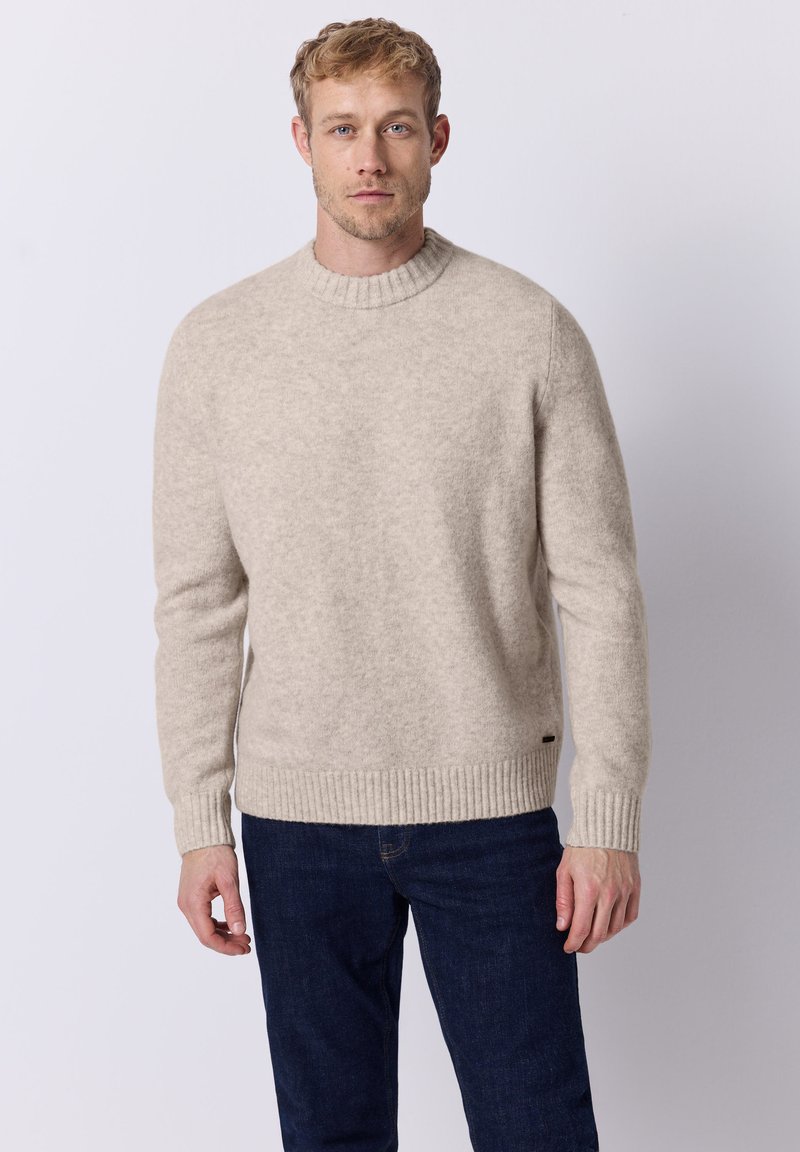 Beige Strickpullover mit geripptem Rundhalsausschnitt und Bündchen, weicher Textur und leicht lockerer Passform, kombiniert mit dunklen Jeans.
