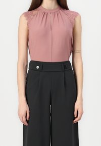 Blouse rose poudré sans manches avec épaules en dentelle, associée à un pantalon noir taille haute à jambes larges avec deux boutons à la taille.