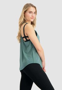 Tanktop aus türkisfarbenem Stoff mit entspannter Passform, ausgestattet mit schwarzen, überkreuzten Trägern auf der Rückseite und einem abgerundeten Saum. Tragen mit schwarzen Leggings.