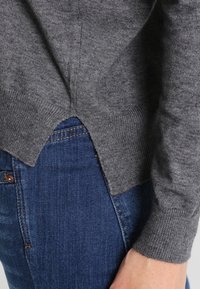 Grau gestrickter Pullover aus weichem Material, mit einem leicht längeren Rücken, gerippten Bündchen und einem seitlichen Schlitz, kombiniert mit blauen Jeans.