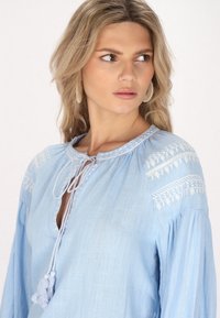 Hellbluse mit weißen bestickten Mustern auf den Schultern, einem V-Ausschnitt mit Bindeschluss und Quasten. Weicher, strukturierter Stoff, lange Ärmel.