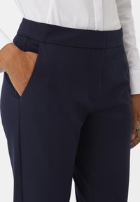 Marineblauwe broek met een gladde textuur, voorzien van een platte tailleband, zijzakken en een getailleerde pasvorm. Close-up van een hand in de zak.