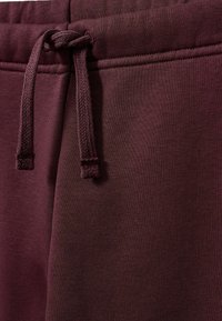 Burgunderfarbene Jogginghose mit strukturiertem Oberflächenmaterial. Sie verfügt über einen Kordelzugbund und kontrastierende Nähte aus einem glatten Stoff.