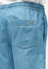 Pantalon en denim bleu clair avec taille élastique, poche arrière ornée d'un petit logo crocodile vert brodé dans le coin supérieur droit.