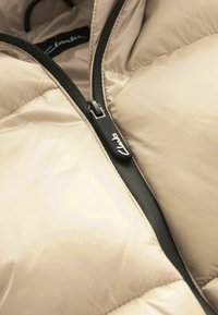 Veste matelassée beige en gros plan montrant une fermeture éclair noire partiellement fermée avec le logo « Clarks » sur la languette.