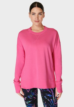 Vrouw die een felroze sweatshirt met lange mouwen en zwarte leggings met kleurrijke abstracte patronen draagt, staand tegen een effen achtergrond.