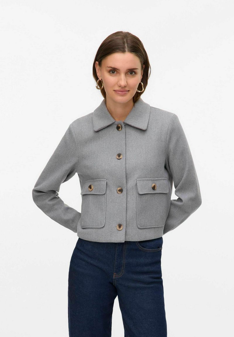 Chaqueta corta de mezcla de lana gris con dos bolsillos frontales, cierres de botones marrones y un cuello clásico, llevada sobre un denim oscuro.