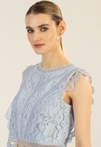 Helleblaue Spitzenbluse mit gewellten Kanten, Blumenmustern und hohem Kragen. Merkmale sind filigrane Spitzenverzierungen und ärmelloses Design.