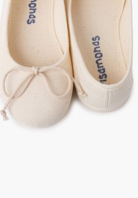 Ballerines en toile beige avec un bout rond, une fine bordure sur les côtés et un nœud décoratif à l'avant. L'intérieur présente une marque bleue sur un tissu clair.