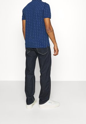 Mężczyzna stojący tyłem, ubrany w niebieską koszulkę polo w wzory, ciemne dżinsy i białe trampki na jednolitym białym tle.