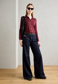 Camisa de botones burdeos de manga larga combinada con pantalones vaqueros de pierna ancha azul oscuro. Un cinturón negro y gafas de sol complementan el conjunto. Texturas suaves.