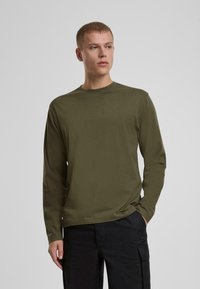 Majica dugih rukava s crewneck izrezom u maslinastoj boji, napravljena od mekanog materijala s opuštenim krojem, s minimalnim šavovima i glatkom teksturom.