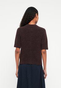 Mulher com cabelo encaracolado vestindo um suéter felpudo de mangas curtas em castanho escuro e uma saia plissada azul-marinho, vista de costas contra um fundo simples.