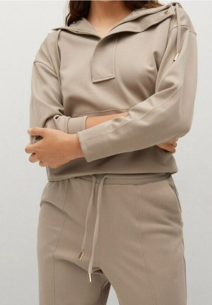 Vrouw draagt beige cropped hoodie met lange mouwen en ritsdetail en bijpassende broek met trekkoord, armen gekruist voor zich.