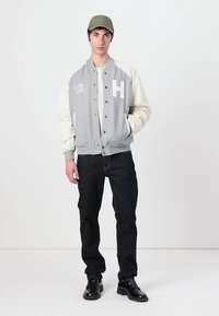 Sivá a krémová bomber bunda s logom "H", s patentkami a bočnými vreckami, kombinovaná s tmavými džínsy a čiernymi topánkami. Zelená čiapka.