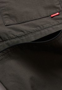 Tmavě zelená tkanina s mřížkovaným vzorem, která má kapsu na zip a červenou štítkovou etiketu. Materiál vypadá hladce a má texturu.