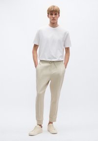 Valkoinen lyhythihainen t-paita yhdistettynä beigenvärisiin collegehousuihin, joissa on kiristysnyöri vyötäröllä ja sivuraidat, viimeistelty beigenvärisillä lenkkareilla.