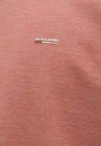 Textúrovaná, svetlooranžová látka s malým strieborným logom "JACK & JONES". Detailný pohľad zblízka zameraný na materiál a detaily značky.