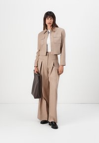 Giacca cropped beige a righe sottili sopra una maglietta bianca, abbinata a pantaloni larghi a righe sottili. La modella tiene una borsa a tracolla di colore grigio scuro. Scarpe nere.