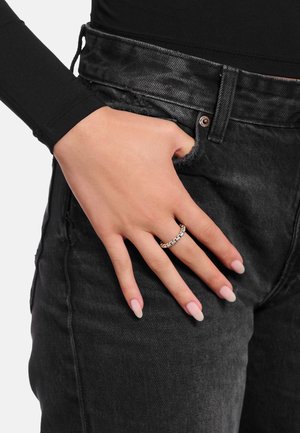 Mano con anello a maglie di catena in argento appoggiata sulla tasca di jeans neri in denim, indossa un top nero a maniche lunghe con unghie a mandorla rosa chiaro.