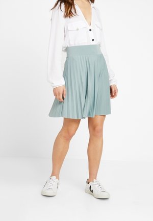 Kvinde iført en hvid bluse med lange ærmer, lyseblåt plisseret nederdel og hvide sneakers med sorte detaljer, stående mod en hvid baggrund.