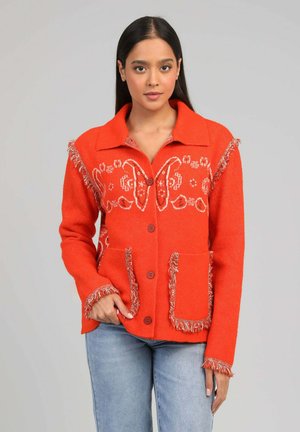 Femme portant un cardigan orange vif à boutons avec des motifs de papillons blancs, des détails à franges, et un jean bleu, sur un fond gris uni.
