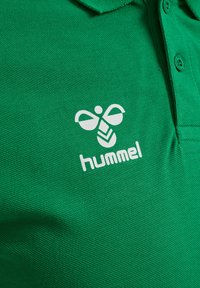 Grön texturerad pikétröja med vit hummel-logotyp och stiliserad biemblem nära bröstknapparna.