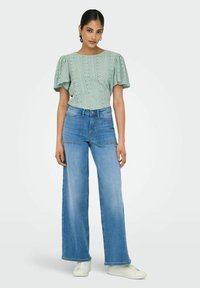 Hellgrünes, strukturiertes Oberteil mit Puffärmeln, kombiniert mit weit geschnittenen blauen Jeans und weißen Sneakers. Dezentes Lochmuster im Stoff.