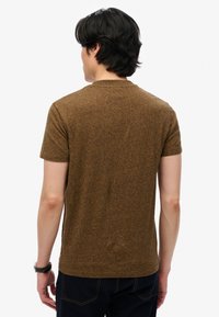 Mand iført en brun kortærmet skjorte og mørke jeans, set bagfra, med sort hår og et ur på venstre håndled.