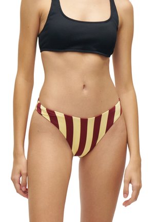 Mujer con un top de bikini negro y braga de bikini de corte alto con rayas verticales burdeos y amarillas, mostrada desde los hombros hasta la parte superior de los muslos.